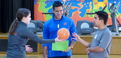 man in blue sweatshirt coaching a young woman and young man passing an orange ball